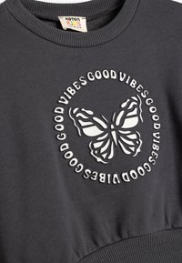 Szara bluza z ribbowanym dekoltem i teksturowaną grafiką motyla. Napis "Good Vibes" otacza motyla w kontrastowej białej haftowanej czcionce.