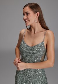 Abito verde scintillante con scollo a cappuccio, spalline sottili e un design testurizzato di paillettes. I pannelli laterali aggiungono contrasto e profondità al tessuto.