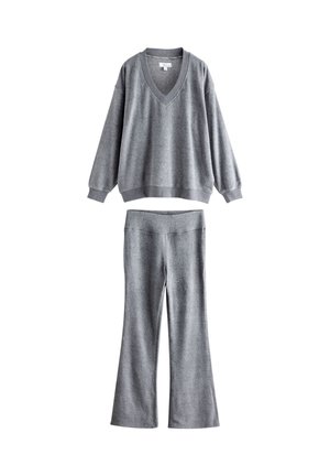 Kvinde står indendørs iført en løs grå sweater, matchende grå bukser og lyserøde slippers på et trægulv nær en pejs.