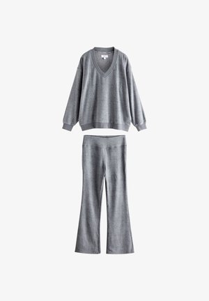 Femeie stând în interior, purtând un pulover gri larg, pantaloni gri asortați și papuci roz pe un podea de lemn lângă un șemineu.