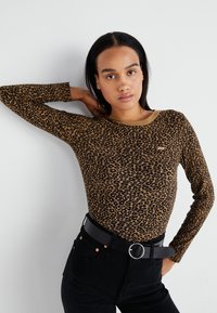 Dugih rukava majica u leopard uzorku s teksturiranom, mekanom tkaninom. Ima okrugli izrez i kombinira se s crnim trapericama visokog struka i crnim remenom.