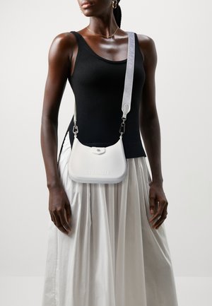 Nainen mustassa topissa ja valkoisessa hameessa, kantaa valkoista olkalaukkua, jossa on leveä hihna olkapäällä.