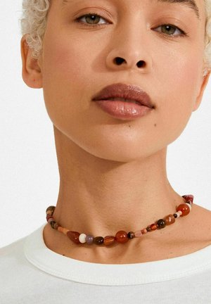 Collier multicolore de perles présentant différentes formes, notamment rondes et en forme de larme, dans des tons de rouge, marron et violet, porté sur un cou visible.