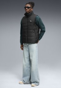 Sort puffer vest med høj krave, lynlåslommer, båret over en grøn langærmet skjorte, parret med lyseblå brede bukser og beige sko.
