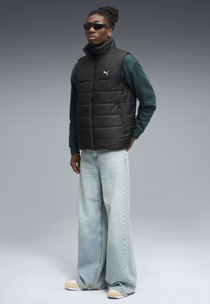 Black puffer vest with high collar, zippered pockets, worn over a green long-sleeve shirt, paired with light blue wide-leg jeans and beige shoes.