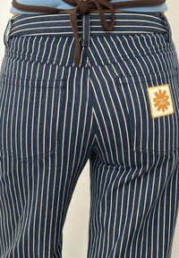 Pantalon bleu marine avec des rayures verticales blanches, poches arrière, taille à cordon marron et un écusson beige sur la poche droite.