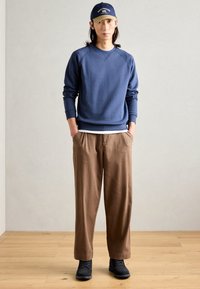 Sudadera de la marina con puños acanalados, pantalones marrones relajados y zapatos negros. Una gorra beige con letras en azul marino completa el atuendo.