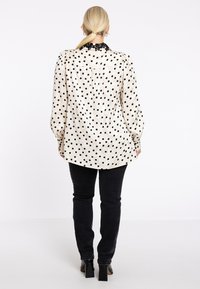 Lange mouw blouse van beige satijn met zwarte stippen; heeft een zwarte kantkraag en een geplooid detail op de rug; gecombineerd met zwarte jeans en hakken.