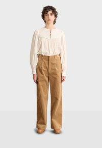Blusa bianca a manica lunga con dettagli di bottoni e ricami, abbinata a pantaloni in corduroy a vita alta e gamba larga in marrone chiaro.
