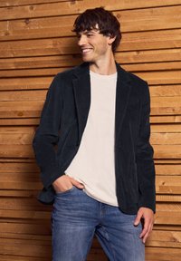 Dark blue corduroy blazer with notch lapels, worn over a light t-shirt, paired with blue jeans. Wooden wall background.