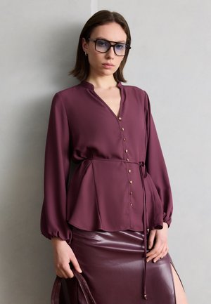 Žena v fialovej blúzke so zapínaním na gombíky a opaskom, zodpovedajúcej lesklej sukni so štrbinou na boku, s veľkými štvorcovými okuliarmi, drží remienok kabelky, stojí opretá o sivú stenu.