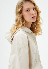 Chaqueta con capucha beige de corte holgado, con costuras visibles y cierre de broche metálico. Textura de tela suave y detalles de diseño casual.