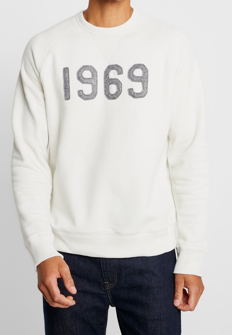 Homme portant un sweat-shirt blanc avec "1969" en chiffres gris texturés, associé à un jean bleu foncé, visible du cou aux hanches.
