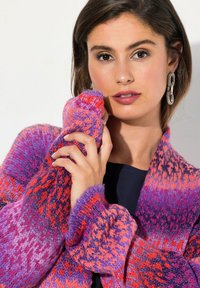 Strickjacke mit einem lebendigen Farbverlauf in Rosa, Lila und Rot. Die Ärmel sind übergroß und weisen ein strukturiertes Muster auf.
