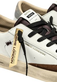 Sneakers bianchi in pelle con accenti scuri, stringhe nere e un'etichetta dorata. Presentano un pannello in suede marrone testurizzato e dettagli del logo.