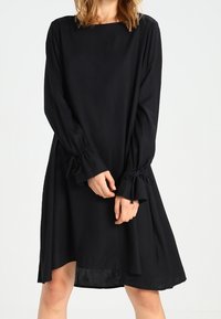 Robe noire à manches longues avec un décolleté carré et des détails froncés au niveau des poignets. Silhouette évasée, longueur genou, fabriquée à partir d'un tissu lisse.