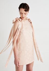 Frau, die ein ärmelloses, hellrosa Jacquardkleid mit floralen Mustern und großen, gebundenen Schleifen auf den Schultern mit langen, herabhängenden Bändern trägt.