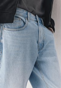 Persoon draagt lichtblauwe spijkerbroek en een zwart jasje, met handen in de voorzakken gestopt, staand tegen een lichte muur.