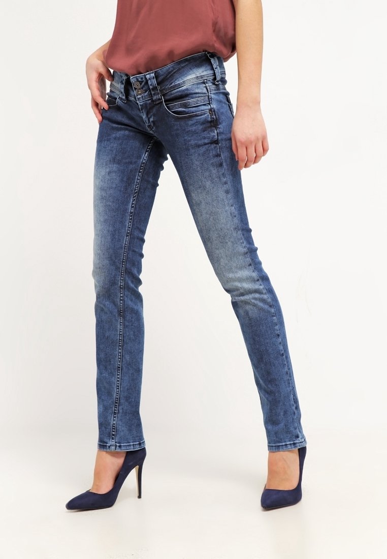 Kvinna iklädd blekta blå tajta jeans och navyblå klackskor, stående mot en enfärgad vit bakgrund.