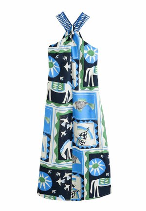 Femme portant une robe dos-nu bleue, noire et blanche avec des motifs abstraits, debout sur une falaise surplombant une mer calme et une côte rocheuse.