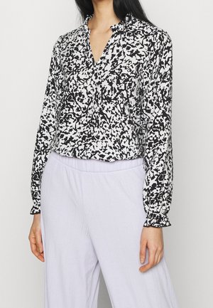 Woman wearing a black and white abstract patterned long-sleeve blouse with ruffled cuffs and light-colored loose pants.
