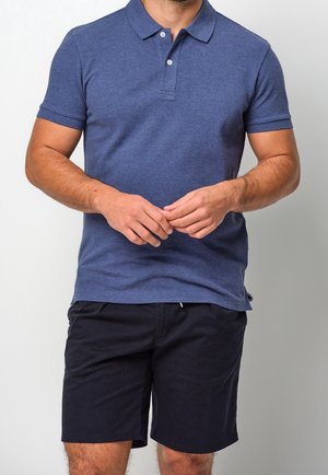 Homme portant un polo bleu à manches courtes et un short noir, debout les mains jointes devant un fond uni.