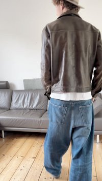 Braune Lederjacke, lockere blaue Jeanshose mit Taschen und ein helles Hemd. Im Hintergrund sind ein graues Sofa und ein Holzboden zu sehen.