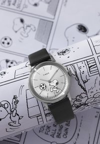 Ρολόι χειρός Timex με μαύρο δερμάτινο λουράκι, που απεικονίζει τον Snoopy να παίζει ποδόσφαιρο σε λευκό καντράν, τοποθετημένο πάνω σε φόντο από κόμικς Peanuts.
