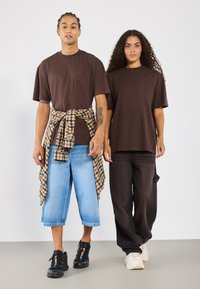 Camisetas de cuello redondo marrones con bolsillos en el frente, culottes de mezclilla azules, pantalones cargo negros. Una persona lleva una camisa de cuadros atada a la cintura.
