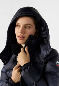 Mujer vestida con una chaqueta acolchada azul marino con la capucha puesta, sosteniendo la cremallera cerca de su barbilla, mirando ligeramente de lado.