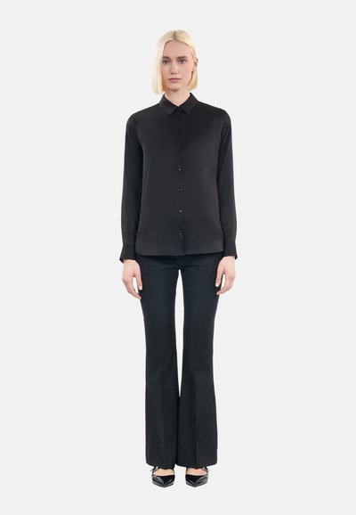 Camicia nera a maniche lunghe con bottoni e colletto, abbinata a pantaloni neri svasati e scarpe nere. Tessuto liscio senza motivi visibili.