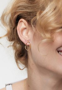 Złote kolczyki w kształcie organicznym noszone przez osobę z kręconymi włosami. Kolczyki mają gładkie, polerowane wykończenie.