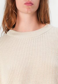 Pull en maille beige clair avec une texture côtelée, doté d'un large col et d'un fit court et décontracté. Gros plan sur le vêtement supérieur.