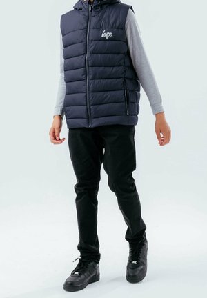 Chaleco acolchado azul marino con capucha, cierre frontal con cremallera y logo en el pecho, combinado con camiseta de manga larga gris y pantalones negros, completado con zapatillas negras.