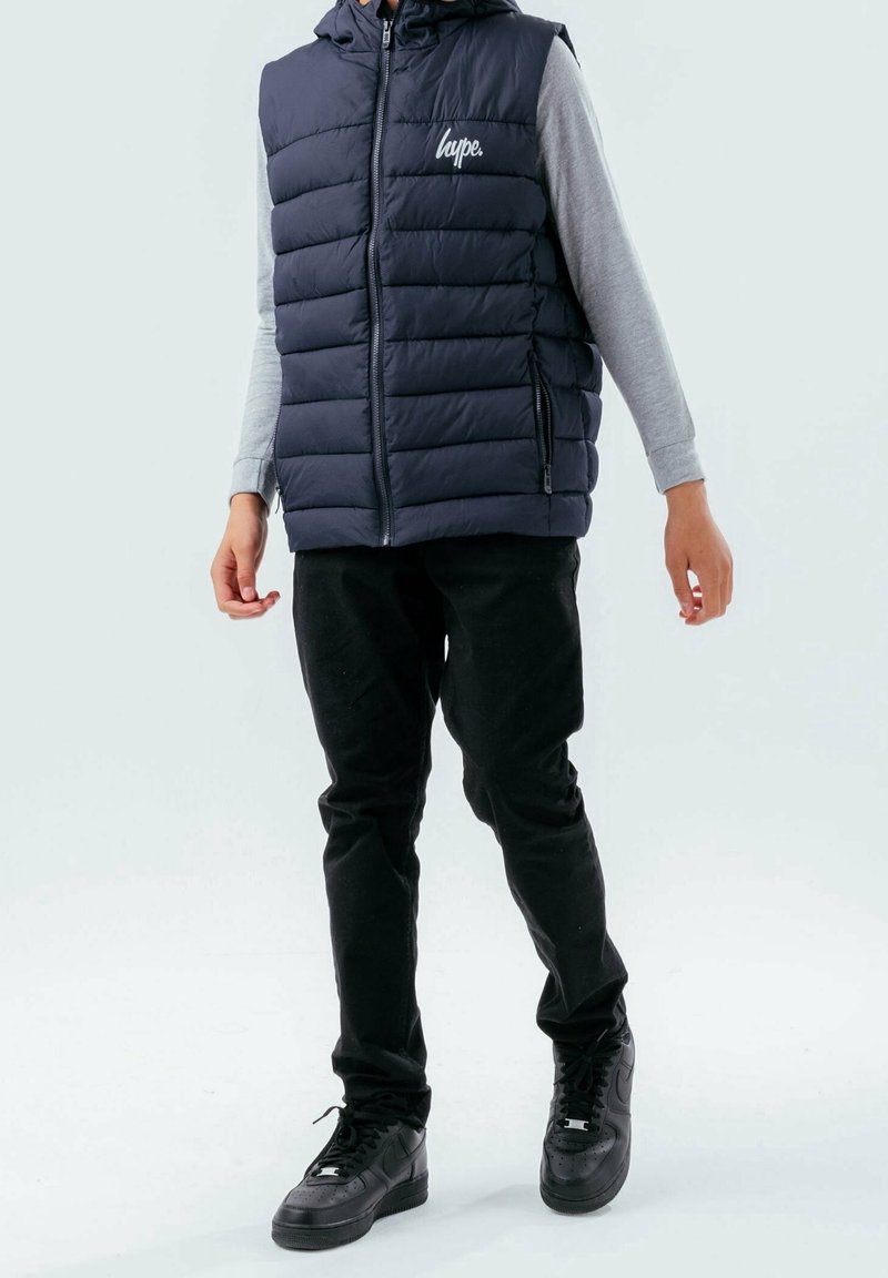 Doudoune sans manches bleu marine avec capuche, fermeture éclair à l'avant et logo sur la poitrine, associée à un haut gris à manches longues et un pantalon noir, complétés par des baskets noires.