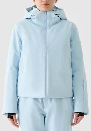 Woman wearing light blue hooded winter jacket and matching pants, with thumbholes in sleeves and front zipper closed.