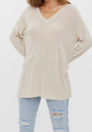 Kobieta w luźnym beżowym swetrze z dekoltem w serek i jasnoniebieskich podartych dżinsach, z rękami założonymi na plecach.