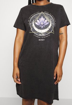 Osoba ubrana w czarną sukienkę typu T-shirt z symbolem lotosu, półksiężyca, trójkąta oraz napisem "Bądź energią, którą chcesz przyciągnąć, Wieczna Siła, MCMXCV".