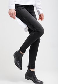 Pantalones vaqueros negros ajustados con una cintura alta y detalles en la costura lateral, combinados con botas de tobillo negras y de punta. Textura de tela suave.