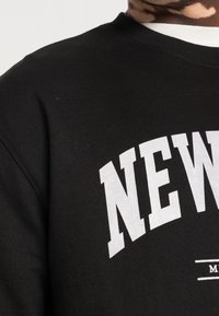 Sudadera negra con texto blanco en negrita que dice "NUEVO" y una línea decorativa. Confeccionada en algodón, tiene una textura suave y un cuello redondo.