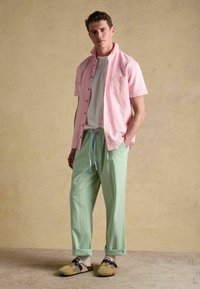 Un jeune homme debout, les mains dans les poches, portant une chemise rose à manches courtes par-dessus un t-shirt blanc, un pantalon rayé vert, des chaussettes et des chaussures à enfiler à motifs.