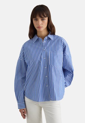 Junge Frau trägt ein blau-weiß gestreiftes Hemd mit Knöpfen und weiße Hose, steht vor einem schlichten hellen Hintergrund.