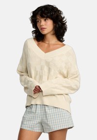 Beige Strickpullover mit kariertem Muster, V-Ausschnitt und übergroßen Ärmeln. Kombiniert mit blauen und weißen karierten Shorts.