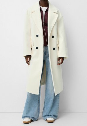 Personne portant un long manteau croisé de couleur blanc cassé, une veste bordeaux à fermeture éclair, un jean large bleu clair et des baskets blanches.