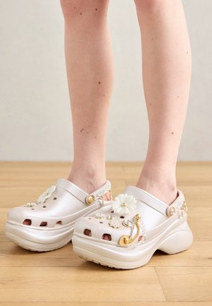 Jambes portant des sabots à plateforme blanc perle décorés de fleurs blanches et de breloques dorées, debout sur un sol en bois.