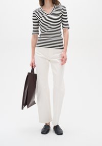 Frau trägt ein schwarz-weiß gestreiftes V-Ausschnitt-Shirt, cremefarbene weite Hosen, schwarze Slipper und hält eine dunkelbraune Umhängetasche.