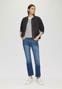 Zwarte bomberjack, gestreept t-shirt met blauwe tekst, blauwe jeans en witte sneakers. De outfit heeft een casual, getailleerde stijl.