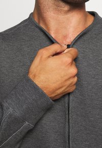 Haut gris à fermeture éclair avec un tissu texturé. Gros plan d'une main saisissant la fermeture éclair. Comprend un col montant et des détails de couture visibles.