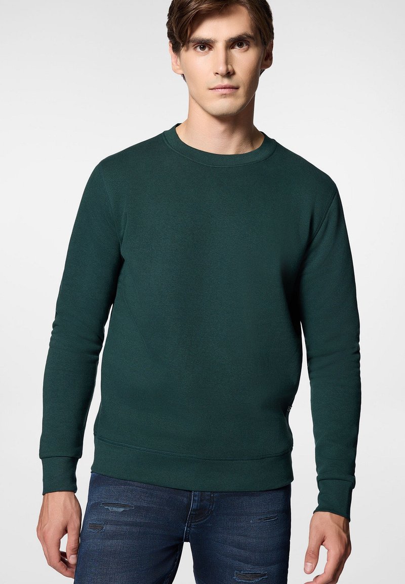 Zielona bluza wykonana z miękkiego materiału, z okrągłym dekoltem, długimi rękawami i ściągaczami. Noszona z ciemnymi podartymi dżinsami.