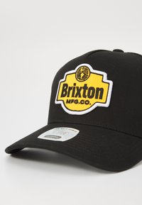Black baseball cap with a yellow and white embroidered Brixton MFG. Co. logo patch. Features a curved brim and ventilation holes.
